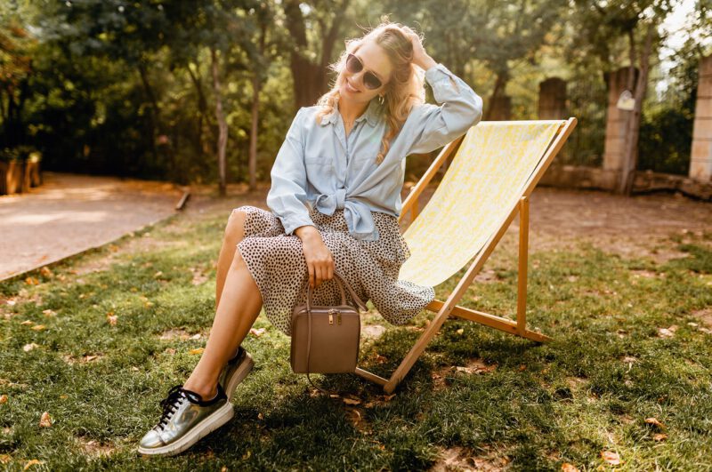 - attractive blond smiling woman sitting deck chair summer outfit blue shirt skirt wearing silver sneakers elegant sunglasses purse street fashion style - ภาพที่ 3